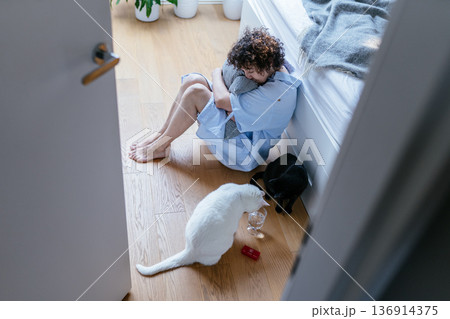 Mature adult female on floor beside bed, hugging pillow tightly, closed defensive posture, glass of water and medication on floor, black and white cats nearby, hormonal mood instability,sleep 136914375