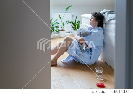 Mature adult female with curly hair and glasses sitting on floor near bed, hugging pillow, glass of water and antidepressants on floor, apathetic expression, hormonal mood changes, insomnia. Mature adult female with curly hair and glasses sitting on floor near bed, hugging pillow, glass of water and antidepressants on floor, apathetic expression, hormonal mood changes, insomnia. 136914385
