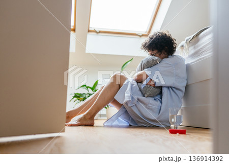 Despaired woman with curly hair and glasses sitting barefoot on bedroom floor near bed, tightly hugging pillow, glass of water and antidepressants, mental health crisis at home 136914392