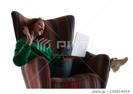 Smiling woman waving during laptop video call in striped armchair. Smiling woman waving during laptop video call in striped armchair. 136914454