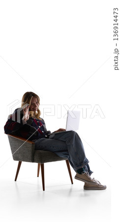 Smiling woman waving during video call on laptop while sitting in armchair isolated on white. Smiling woman waving during video call on laptop while sitting in armchair isolated on white. 136914473
