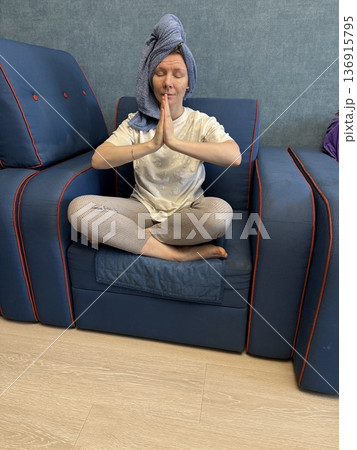 A woman with a blue towel on her head sits on a blue sofa with hands in a prayer pose, morning wellness, meditation practice, home spa. A woman with a blue towel on her head sits on a blue sofa with hands in a prayer pose, morning wellness, meditation practice, home spa. 136915795