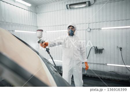 An auto painter in a protective suit is skillfully spraying a car in a paint booth An auto painter in a protective suit is skillfully spraying a car in a paint booth 136916399