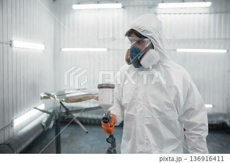 A worker dressed in a protective suit is spray-painting a car panel inside a modern paint booth 136916411