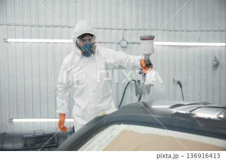 Automotive technician spray-painting car panel in protective suit inside paint booth 136916413