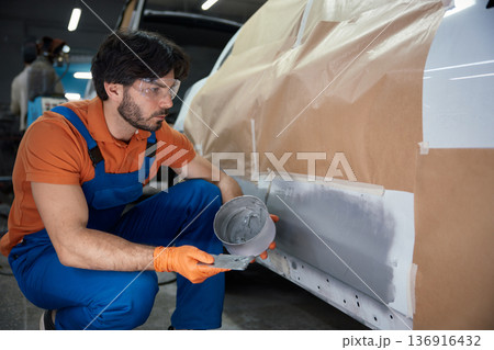 Technician is meticulously prepping a car panel with primer in a garage workshop 136916432