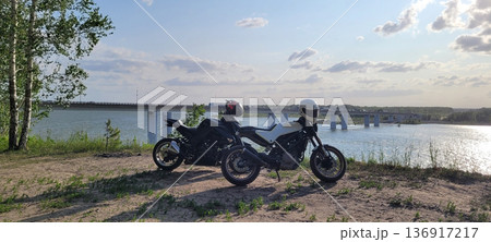 Two motorcycles with helmets parked on sandy ground near a river and a bridge. Travel and adventure concept for motorcycling lifestyle. 136917217