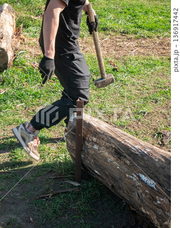 Man in black clothes holding a sledgehammer, standing near a wooden log on the ground. Outdoor activity, manual labor. Man in black clothes holding a sledgehammer, standing near a wooden log on the ground. Outdoor activity, manual labor. 136917442