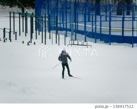 Man skiing on a snow covered field next to playground equipment and a blue fence. Winter sports and outdoor recreation for healthy lifestyle. 136917522