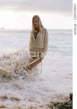 Blonde woman in flowing beige dress and splashing sea waves. 136919607