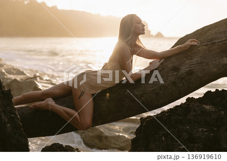 Fashionable woman in a dress posing on wooden log with sunset tones 136919610