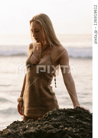 Blonde woman in dress standing on rocks by sea at sunset 136919612