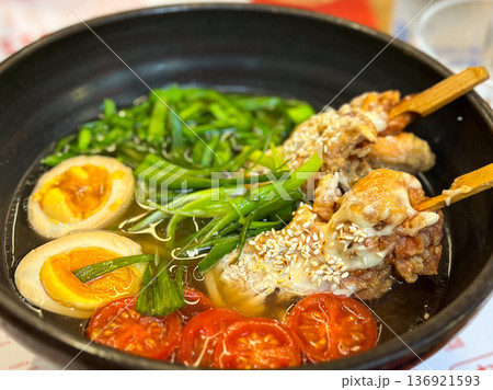 Closeup ramen bowl with vegetables, egg and chicken skewer on wooden table in restaurant. Comfort food and cultural dining experience Closeup ramen bowl with vegetables, egg and chicken skewer on wooden table in restaurant. Comfort food and cultural dining experience 136921593