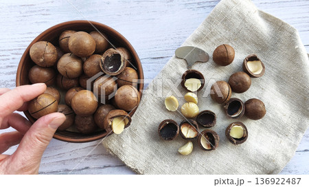Macadamia nuts in a bowl with cracked shells and kernels on linen cloth. Hand picking nut, nutcracker tool nearby. Natural organic snack preparation, rustic flat lay food background. 136922487