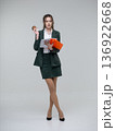 Happy young business woman in a dark green suit standing full length on a white background, holding an orange folder with papers and a coffee cup, looking away. Curly hair, brown-haired 136922668