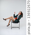 An elegant white woman in a green business suit talks on a red telephone while sitting on a chair against the backdrop of an isolated space. Office. Curly hair, brown-haired 136922670