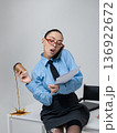 Young white woman in office is talking on the phone holding a cup of coffee and a folder with documents sitting on the edge of a table in business attire Blue shirt glasses Isolated space Pours coffee 136922672