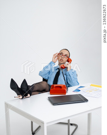 Young calm white woman in office attire working on laptop while talking on vintage red phone, isolated. Business occupation. In a blue shirt with a tie. Freelance . relaxation 136922673
