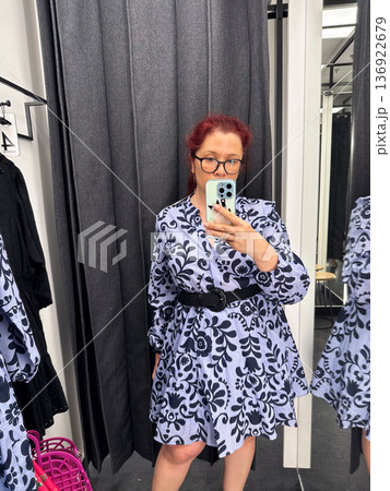 Woman trying patterned dress in fitting room mirror. Fashion choice, retail shopping, style evaluation and modern consumer lifestyle Woman trying patterned dress in fitting room mirror. Fashion choice, retail shopping, style evaluation and modern consumer lifestyle 136922679