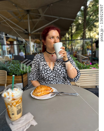 Cheerful millennial woman enjoys a hot beverage and a croissant at an outdoor cafe on a sunny day. Summer moment of joy and tranquility surrounded by lush greenery and urban restaurant charm Cheerful millennial woman enjoys a hot beverage and a croissant at an outdoor cafe on a sunny day. Summer moment of joy and tranquility surrounded by lush greenery and urban restaurant charm 136922697