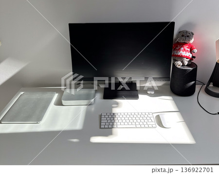 Minimalist white workspace with a computer monitor, keyboard, and other tech accessories on a clean desk. Bright sunlight streaming through the window highlights the neat and organized setting 136922701