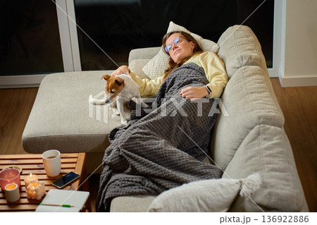 Woman lying on sofa under blanket resting with dog beside her 136922886