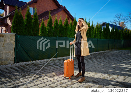 Woman in coat standing with suitcase and looking into distance on street Woman in coat standing with suitcase and looking into distance on street 136922887