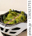 Inside a white bike helmet, a tiny diorama shows three cyclists riding a dirt trail through grassy hills 136923755