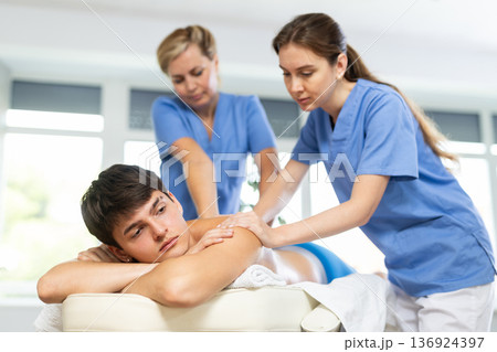 Two female masseuses perform stroking and rubbing of muscles of back and shoulders to guy client 136924397