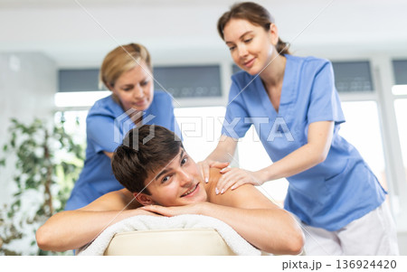 Female professional physiotherapist helps girl student to perform back massage to male client. Female professional physiotherapist helps girl student to perform back massage to male client. 136924420