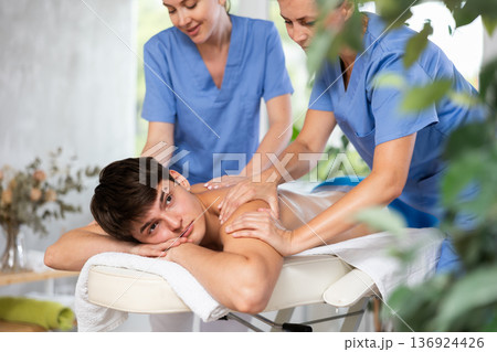 Relaxed young man having back massage therapy conducted by two masseuses Relaxed young man having back massage therapy conducted by two masseuses 136924426