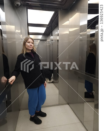 A woman stands inside a modern elevator with metallic mirrored walls, urban lifestyle, daily commute, modern interior. 136925423