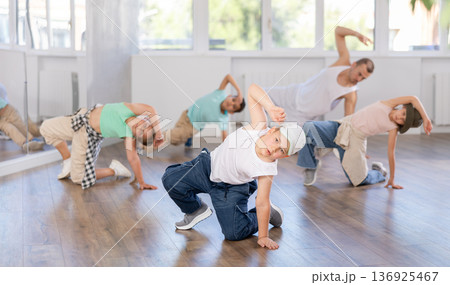 Expressive cheerful teen boy practicing hip-hop movements on floor during group dance class with teacher in studio 136925467