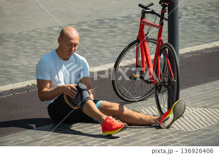 Mature cyclist sitting on pavement holding knee after workout 136926406