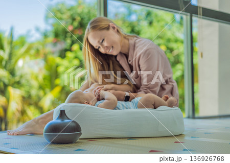 Newborn baby resting in a cozy cocoon beside mother with aroma lamp, calm home atmosphere. Infant relaxation, gentle care, motherhood routine, aromatherapy for babies and family wellbeing concept. Newborn baby resting in a cozy cocoon beside mother with aroma lamp, calm home atmosphere. Infant relaxation, gentle care, motherhood routine, aromatherapy for babies and family wellbeing concept. 136926768