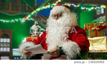 Santa Claus looks at letters received from kids, saluting and welcoming them. Smiling person in Santa costume reads wish lists written by children for Christmas, waving hand, greeting them, camera A Santa Claus looks at letters received from kids, saluting and welcoming them. Smiling person in Santa costume reads wish lists written by children for Christmas, waving hand, greeting them, camera A 136926968