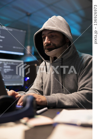 Hooded injured man working diligently on cybersecurity, analyzing networks and spyware risks. Focused male programmer with neck brace writing code and concentrating on improving company networks. 136926973
