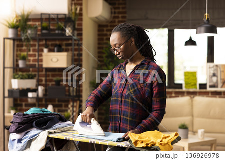 Pretty black woman stands in brick wall living room, ironing wrinkled shirt with electric iron. Calm and focused, african american housewife completes daily household chores at home. 136926978