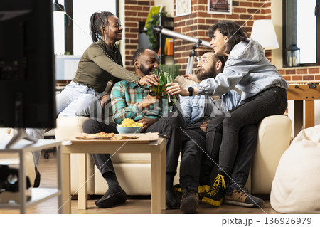 Joyful multiethnic friends celebrate together on couch, raising beer bottles and sharing snacks in cozy brick wall living room. Young people enjoying casual hangout and socializing at home. 136926979