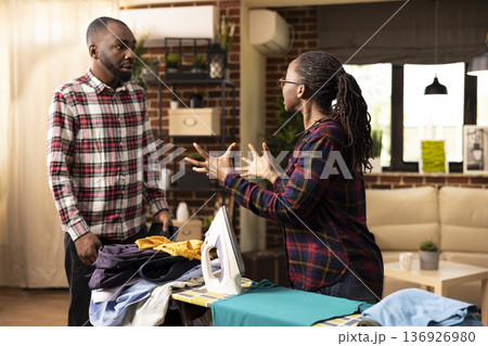 Exhausted black woman takes break from ironing shirts, angrily confronts boyfriend who avoids helping with household chores. Tired lady looks upset, showing tension and frustration over daily 136926980
