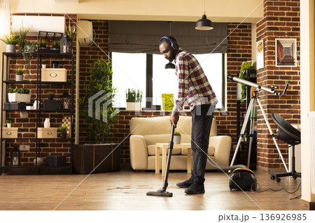 African american homeowner listening to podcast while cleaning brick wall apartment. Young man vacuums hardwood floors in living room, combining chores with entertainment during weekend routine. 136926985