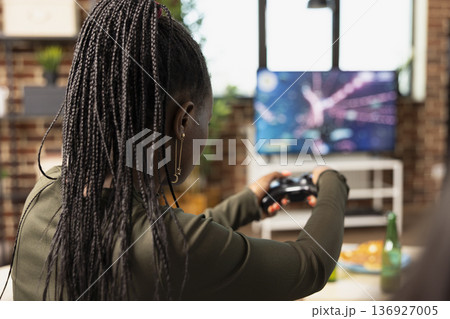 Young black woman plays console game on couch, holding joystick and enjoying fun digital action. Diverse friends sits with wireless controllers, competing in online video game during casual hangout. Young black woman plays console game on couch, holding joystick and enjoying fun digital action. Diverse friends sits with wireless controllers, competing in online video game during casual hangout. 136927005