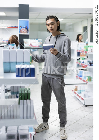 Young adult examines supplement boxes while shopping in medical retail store. Male customer compares product information to balance curiosity with informed choices and mindful healthcare purchasing. Young adult examines supplement boxes while shopping in medical retail store. Male customer compares product information to balance curiosity with informed choices and mindful healthcare purchasing. 136927021