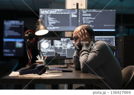 Frustrated software engineer leans forward, holding face in exhaustion and reviewing errors. Tired programmer works late at desk surrounded by coding interfaces and system windows. 136927069
