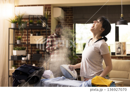 Cheerful woman holding iron with steam, singing and ironing clothes on board. Caucasian female individual enjoys laundry chores at home, working with music and positive energy indoors. 136927070