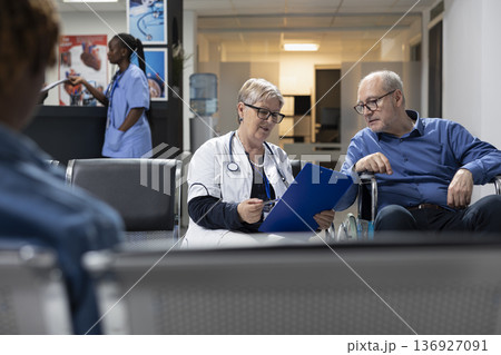 Senior female physician writes medical notes on clipboard while speaking with elderly male patient. Older man in wheelchair listens calmly, receiving guidance and information in hospital lobby. 136927091