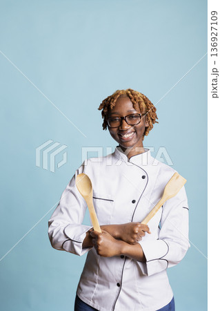 Cheerful black professional cook stands in studio holding wooden cooking utensils, posing with confident and determined expression. Young woman wears white chef uniform against blue background. Cheerful black professional cook stands in studio holding wooden cooking utensils, posing with confident and determined expression. Young woman wears white chef uniform against blue background. 136927109