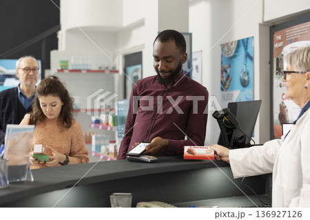 African american man pays for pharmacy items using smartphone, demonstrating cashless payment methods. Senior female pharmacist provides efficient customer service as black customer purchases medicine African american man pays for pharmacy items using smartphone, demonstrating cashless payment methods. Senior female pharmacist provides efficient customer service as black customer purchases medicine 136927126