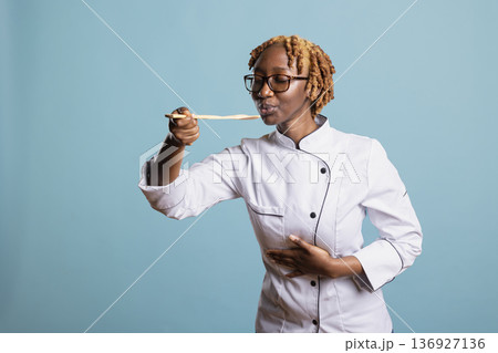 Smiling female chef tastes food from wooden spoon while posing in blue background studio. Professional cook enjoys tasting her creation, expressing satisfaction and passion for culinary craftsmanship. Smiling female chef tastes food from wooden spoon while posing in blue background studio. Professional cook enjoys tasting her creation, expressing satisfaction and passion for culinary craftsmanship. 136927136