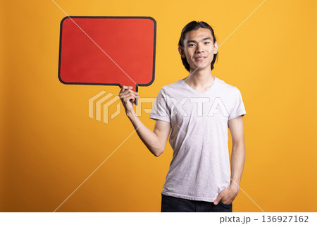 Young asian man stands confidently holding blank placard while posing with one hand in pocket. Focused male model holds signboard, presenting clear copy space for business or promotional ads. Young asian man stands confidently holding blank placard while posing with one hand in pocket. Focused male model holds signboard, presenting clear copy space for business or promotional ads. 136927162